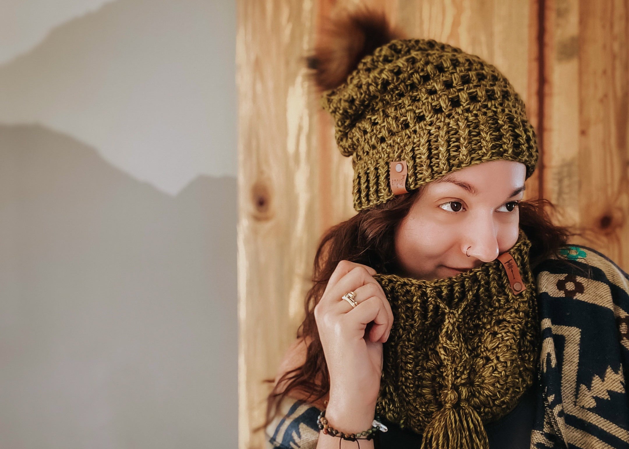 Person wearing a green crochet hat and scarf against a wooden wall.