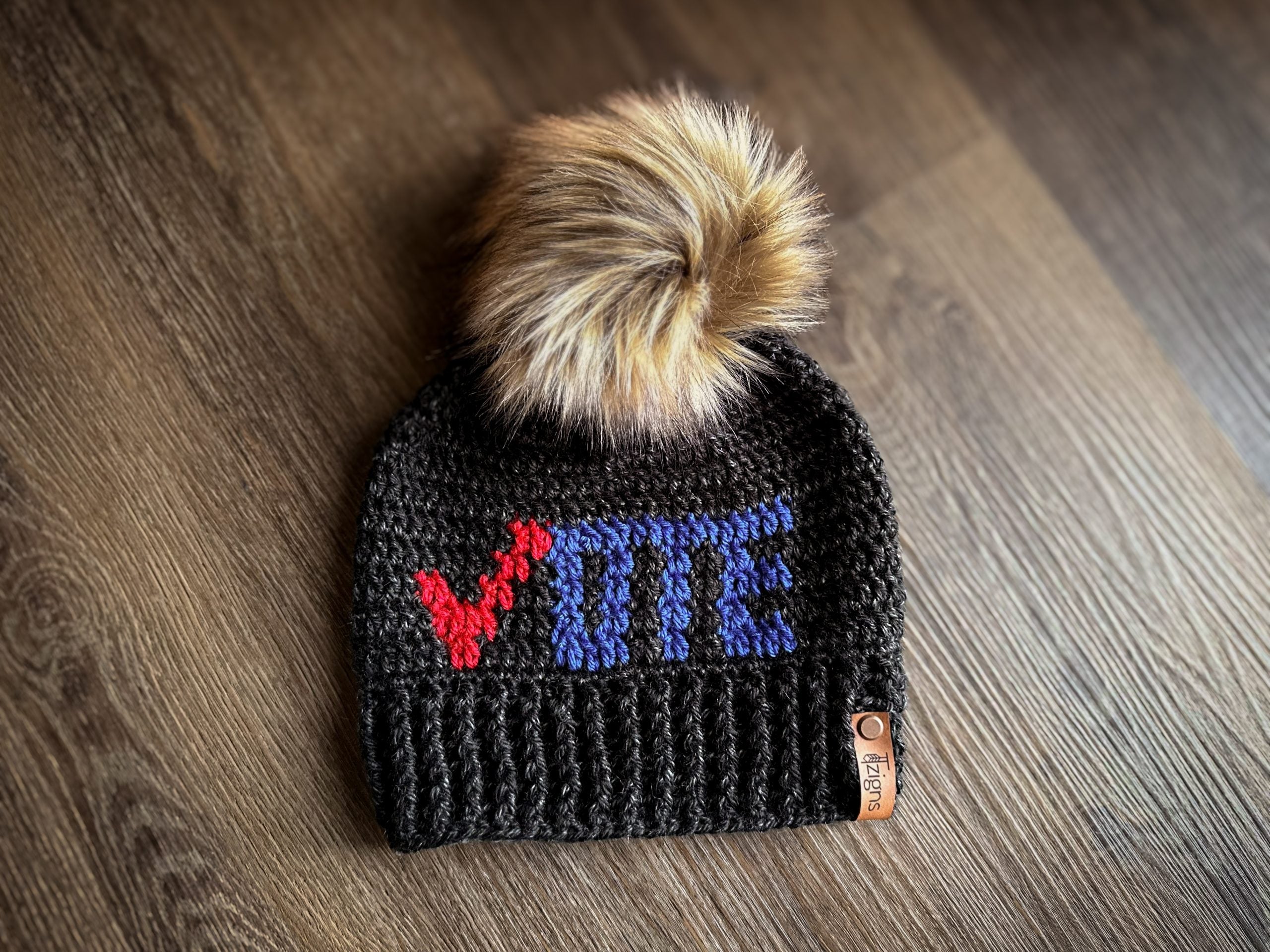 A crochet black hat with the words “vote” on it in red and blue. The hat has a large, light brown faux fur pom pom on it.