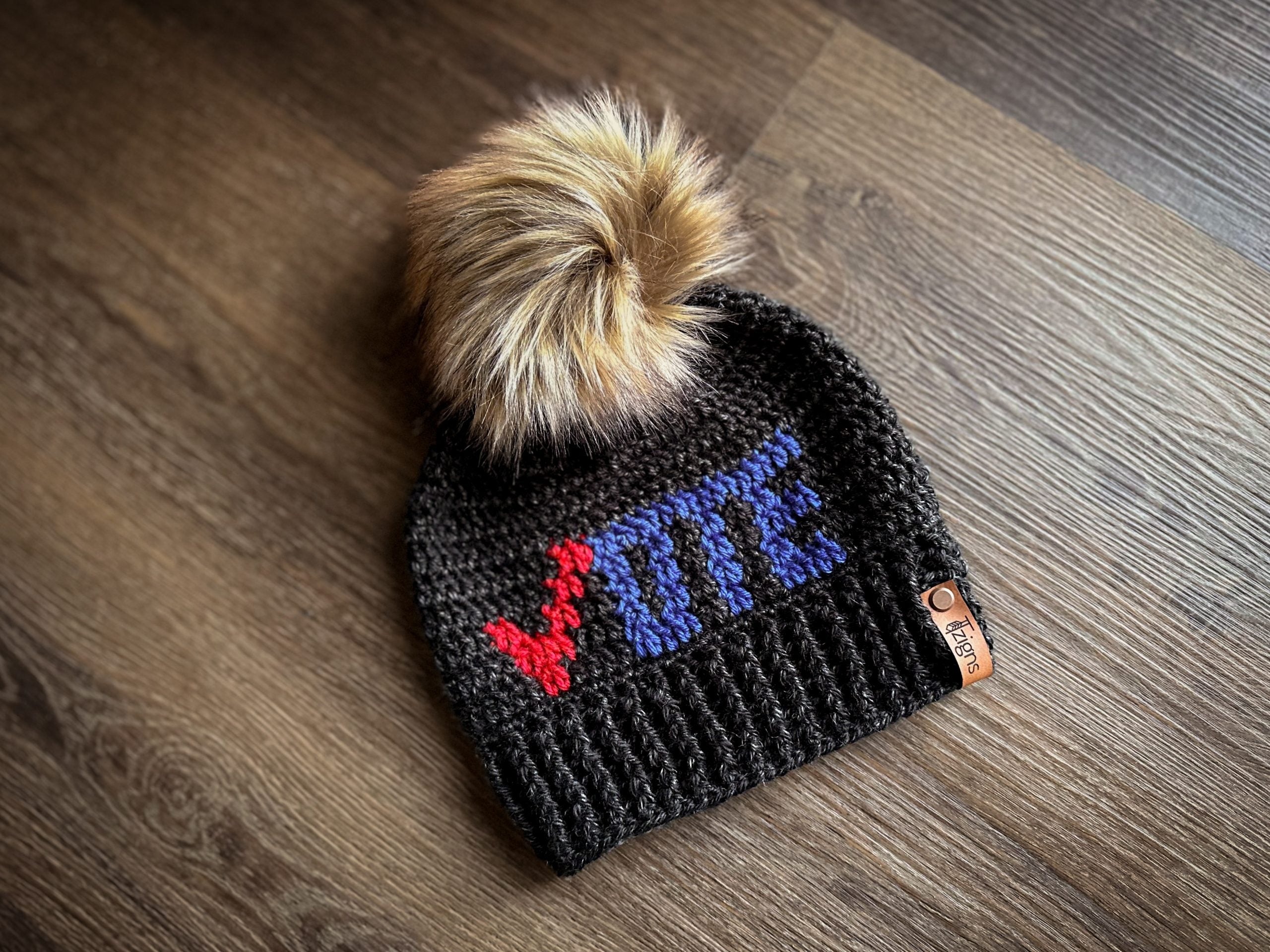 A crochet black hat with the words “vote” on it in red and blue. The hat has a large, light brown faux fur pom pom on it.