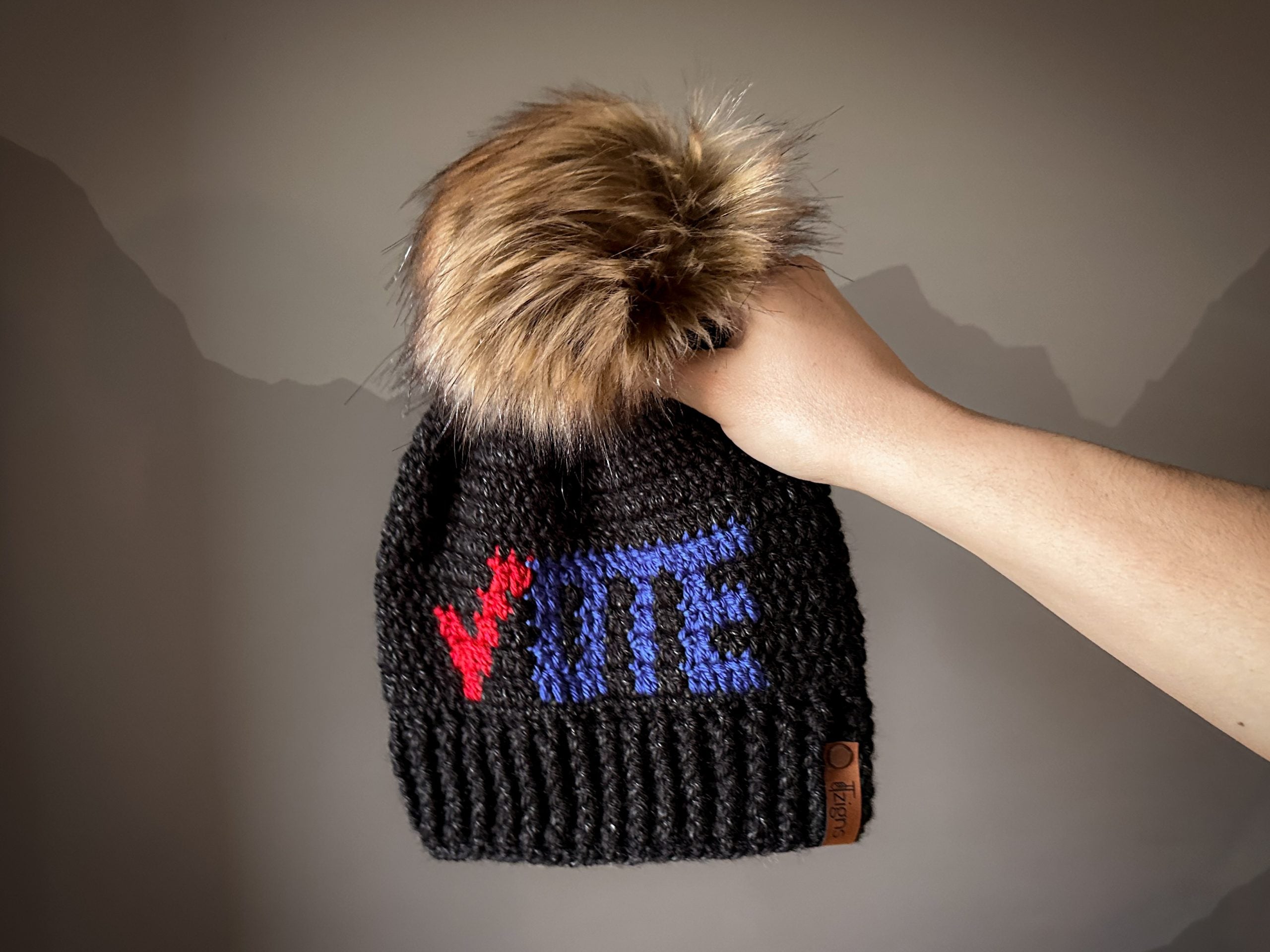 Someone is holding a crochet black hat with the words “vote” on it in red and blue. The hat has a large, light brown faux fur pom pom on it.