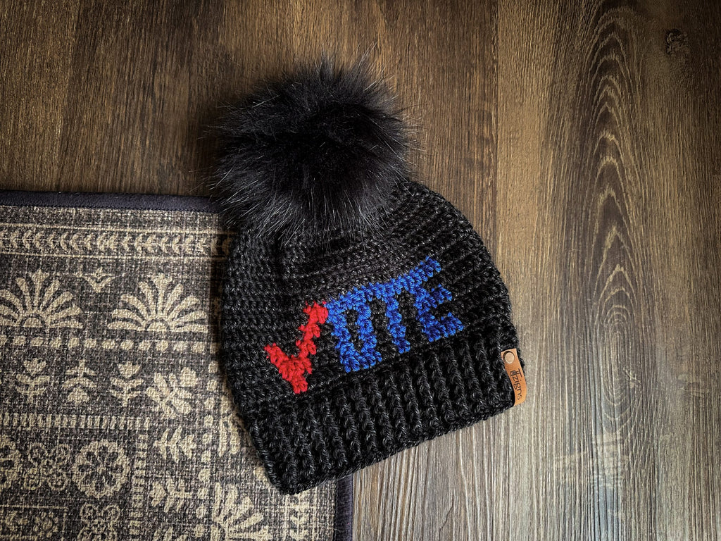 A crochet black hat with the words “vote” on it in red and blue. The hat has a large, black faux fur pom pom on it.
