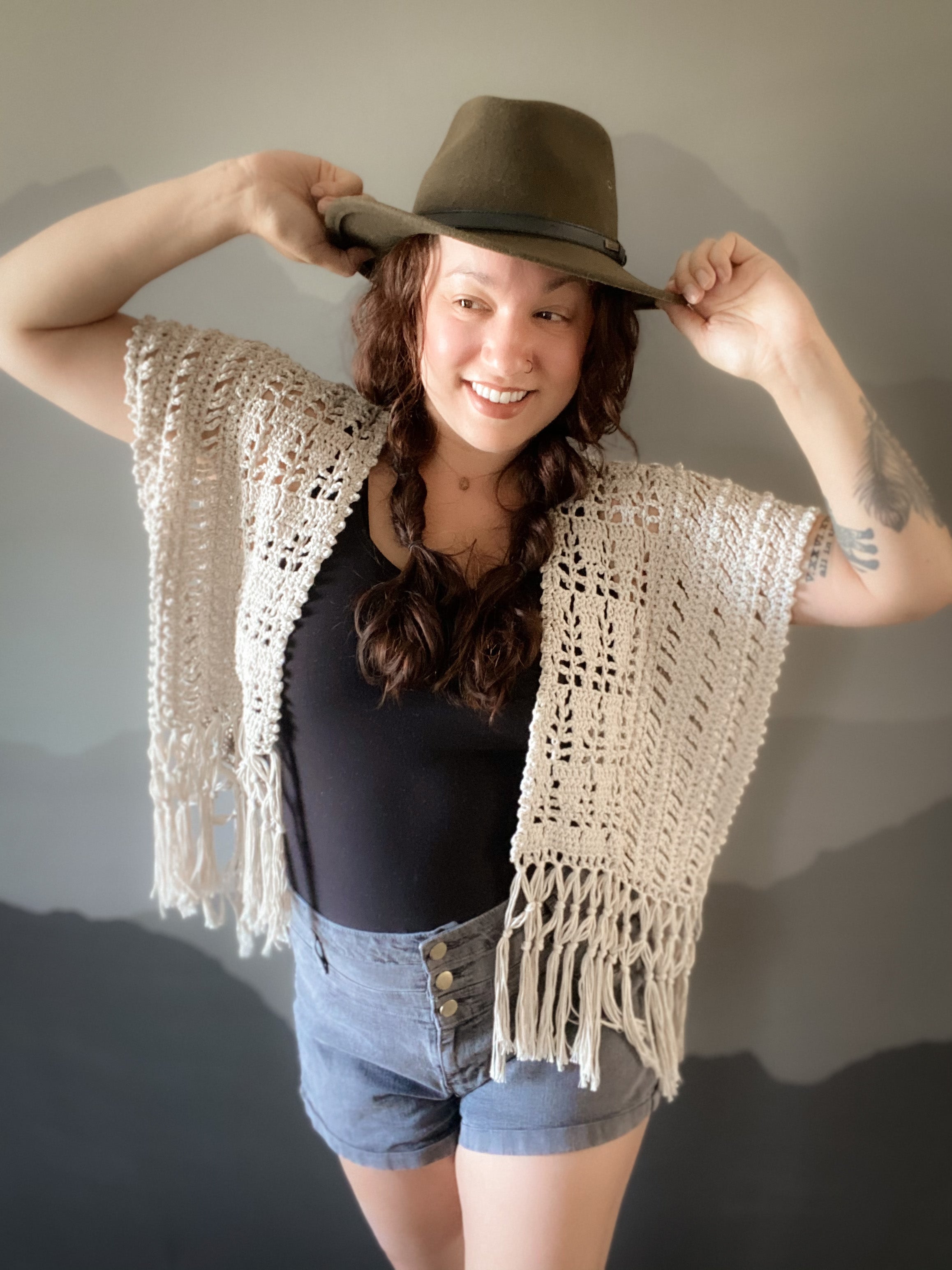 Woman wearing a crochet vest, black top, denim shorts, and a brown hat against a plain background
