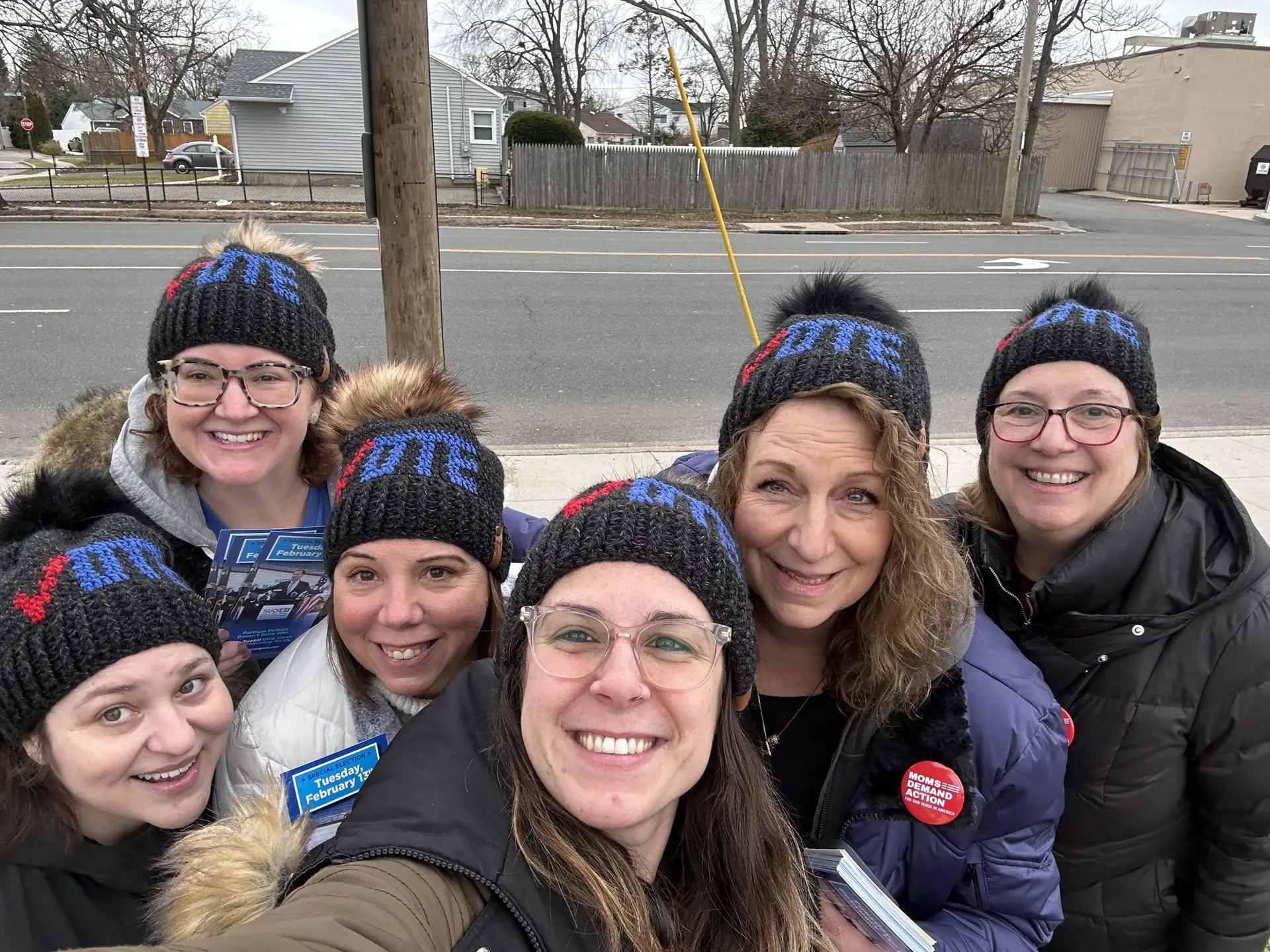 Group of 6 people, outside, wearing handmade crochet black hats with the word Vote in red and blue on them. Hats have black and brown faux fur pom poms on them.