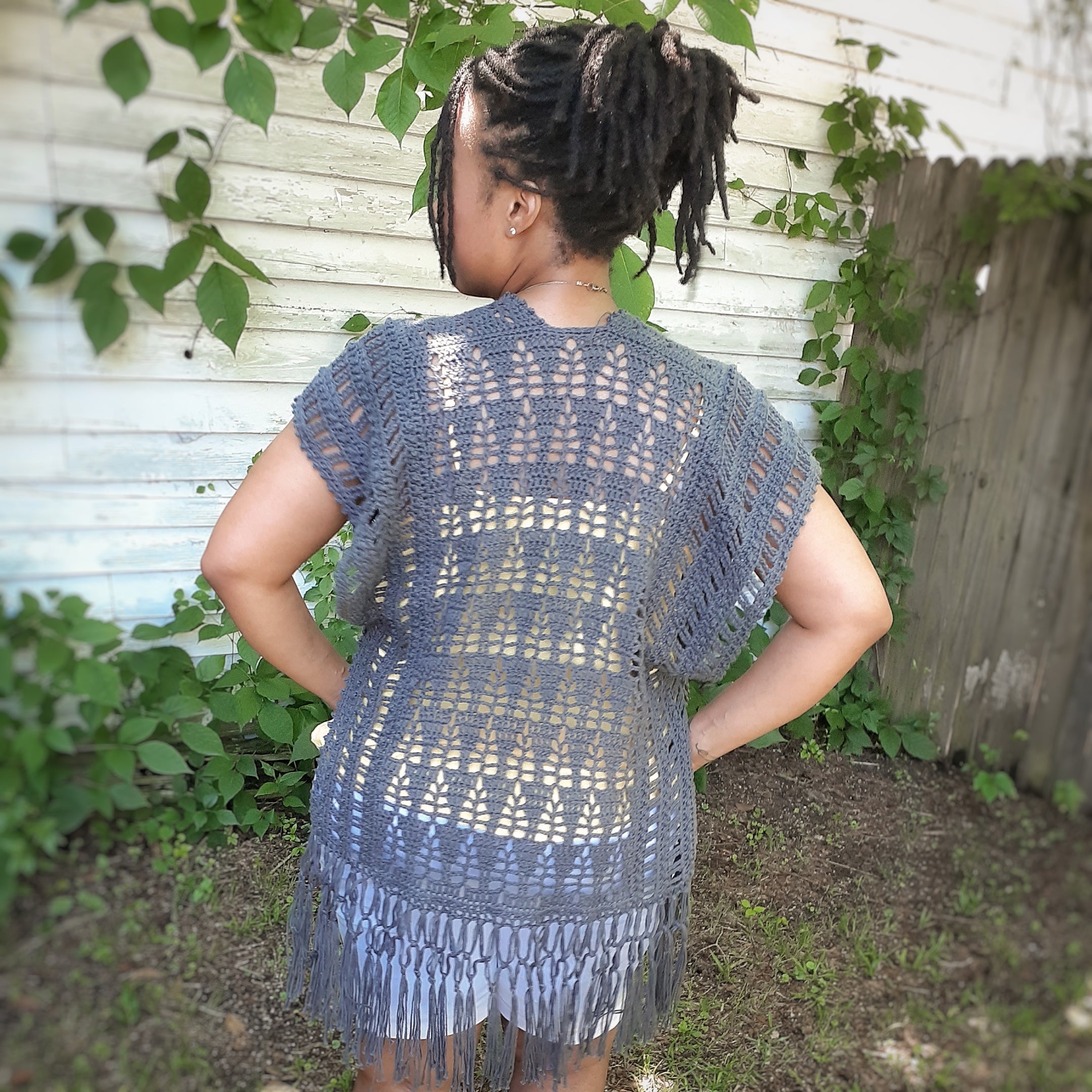 Person wearing a gray crochet cover-up with fringe, standing outdoors against a wooden fence and greenery.