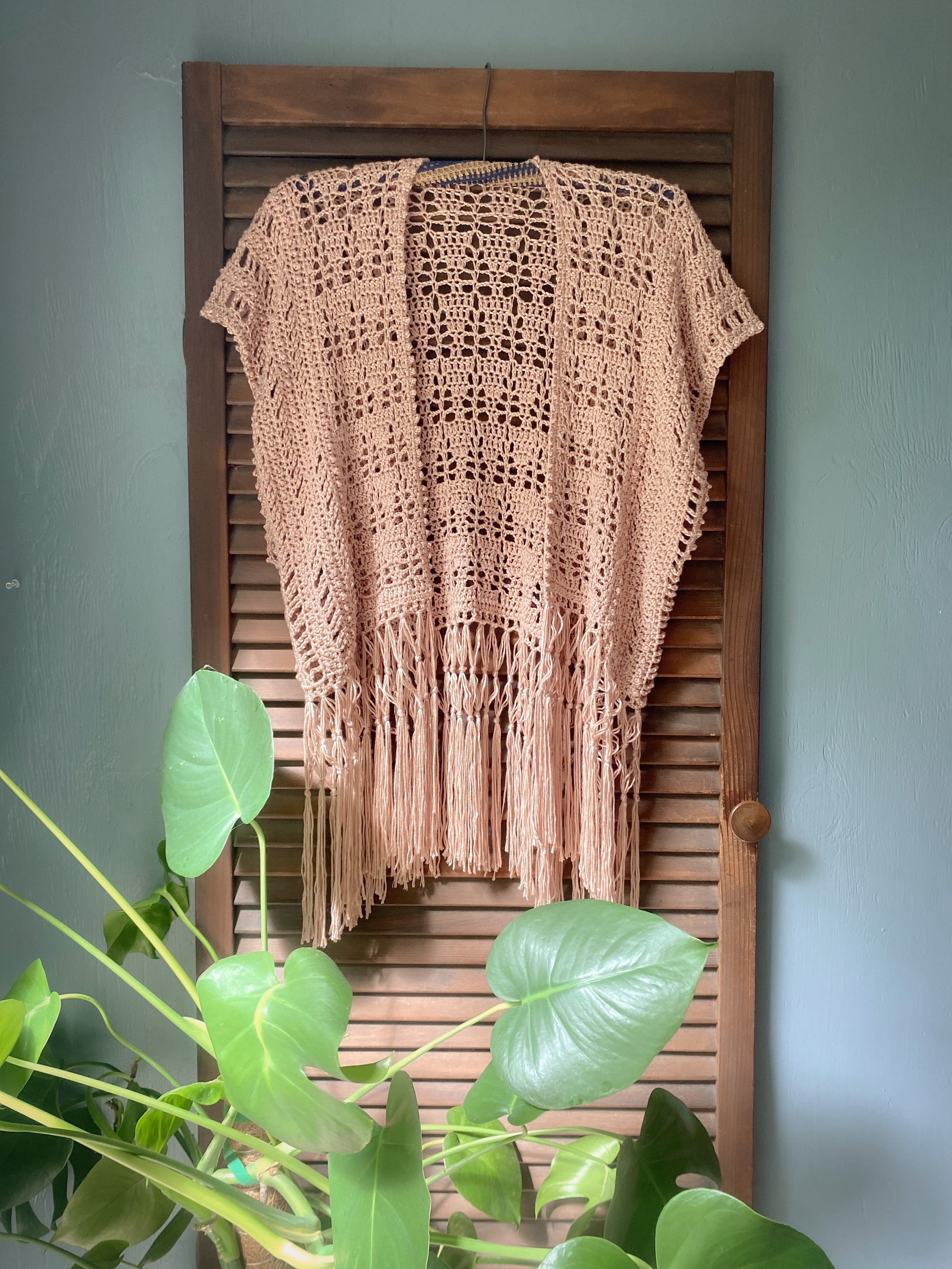 Brown crochet fringe sweater hanging on a wooden shutter with green plants in the foreground.